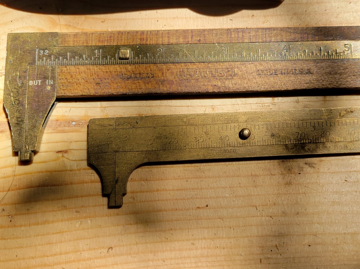 Two old brass sliding calipers on a pine bench, one with inch marks, one with millimetres, beside the heel of a hand plane.