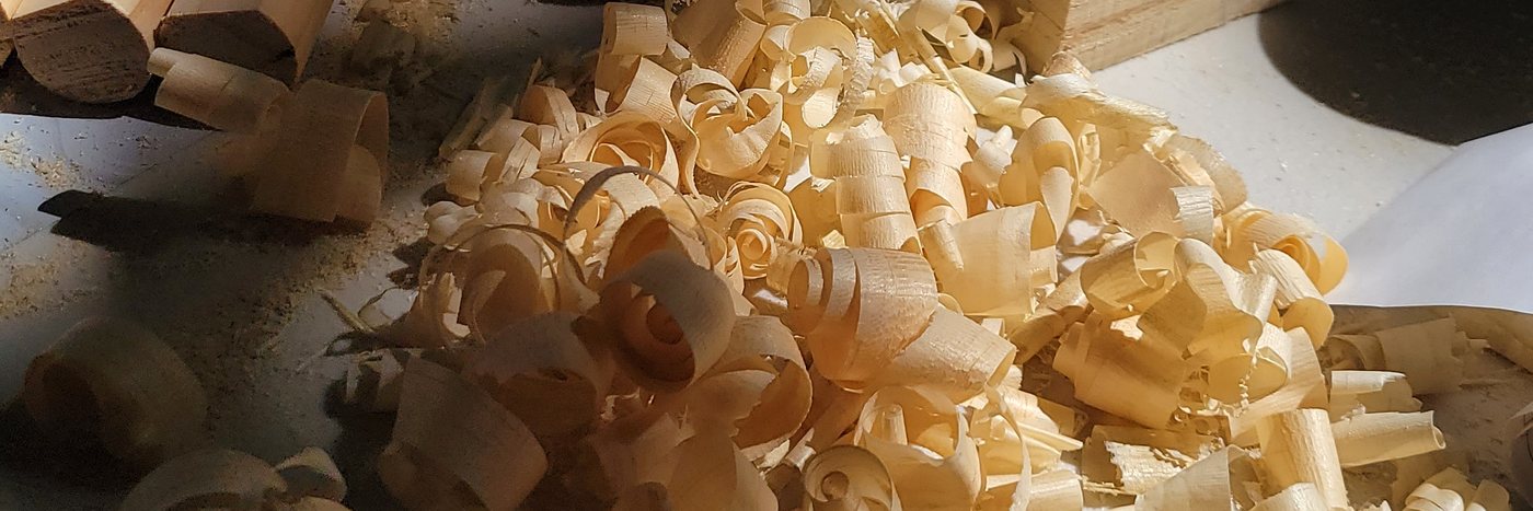 A pile of curled pine shavings in afternoon light, beside freshly cut dowels and blocks on Terry's bench.